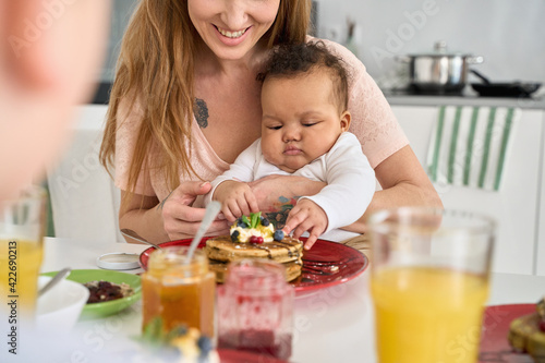 Cute adorable infant mixed race baby child girl sitting on mom lap tasting pancakes at kitchen table. Happy white mother holding little adorable kid daughter having family breakfast at home.