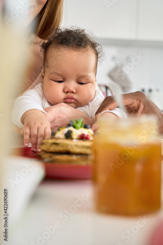 Cute adorable funny infant mixed race baby child girl playing while multiethnic diverse parents having pancakes on family breakfast sitting at kitchen table at home. Close up view