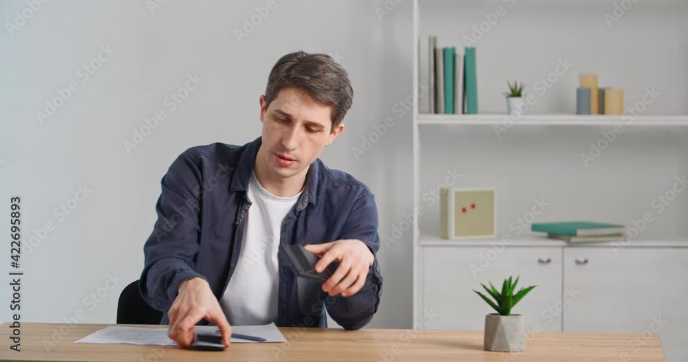 Caucasian tired business man overworked exhausted guy sitting at table, by mistake dials number and calls from calculator, feels confusion talking on mobile phone looking at camera with sad expression Caucasian tired business man overworked exhausted guy sitting at table, by mistake dials number and calls from calculator, feels confusion talking on mobile phone looking at camera with sad expression