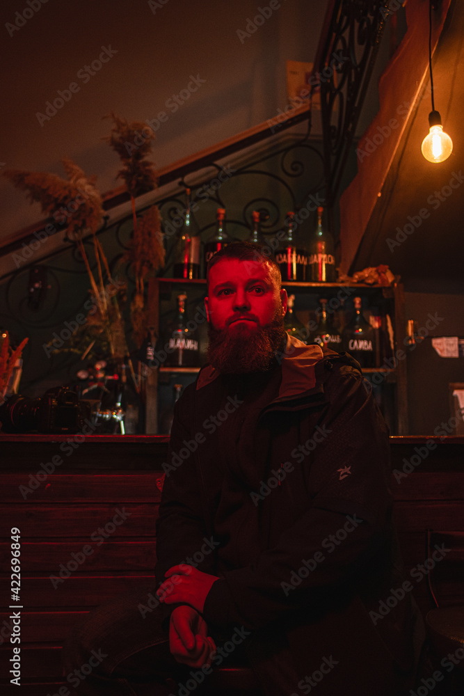 Portrait of a bearded man who sits near a bar, a camera is on the table, red lighting, there is a slight noise, soft focus Portrait of a bearded man who sits near a bar, a camera is on the table, red lighting, there is a slight noise, soft focus