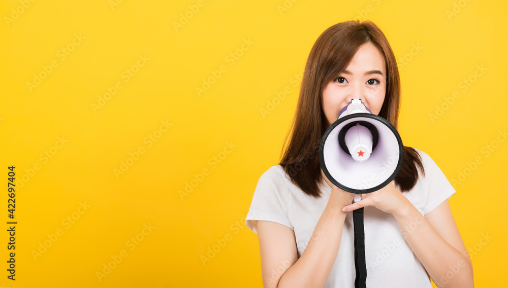 Fototapeta premium Asian happy portrait beautiful cute young woman teen standing making announcement message shouting screaming in megaphone looking to camera isolated, studio shot on yellow background with copy space