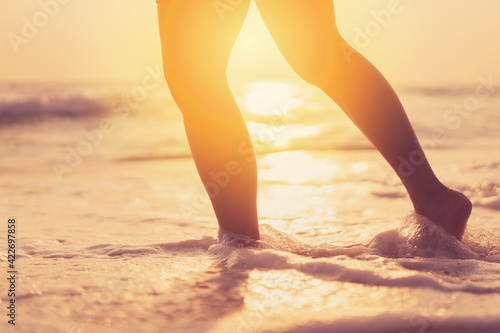 Fototapeta Naklejka Na Ścianę i Meble -  Close up woman legs walking on tropical sunset beach with smooth wave and bokeh sun light wave abstract background. Travel vacation and freedom feel good concept.
