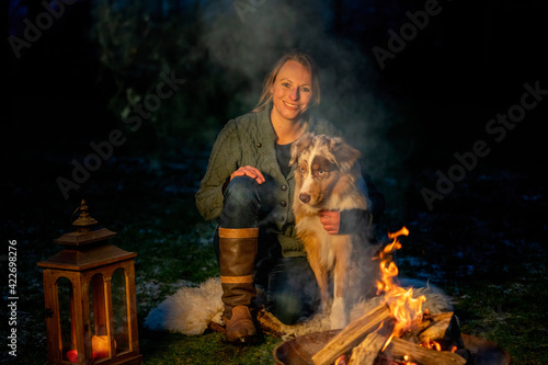 An Australian Shepherd dog sits with his owner in the twilight by the campfire. Woman and dog are lit by the gold colored fire at dusk. Camping life in winter