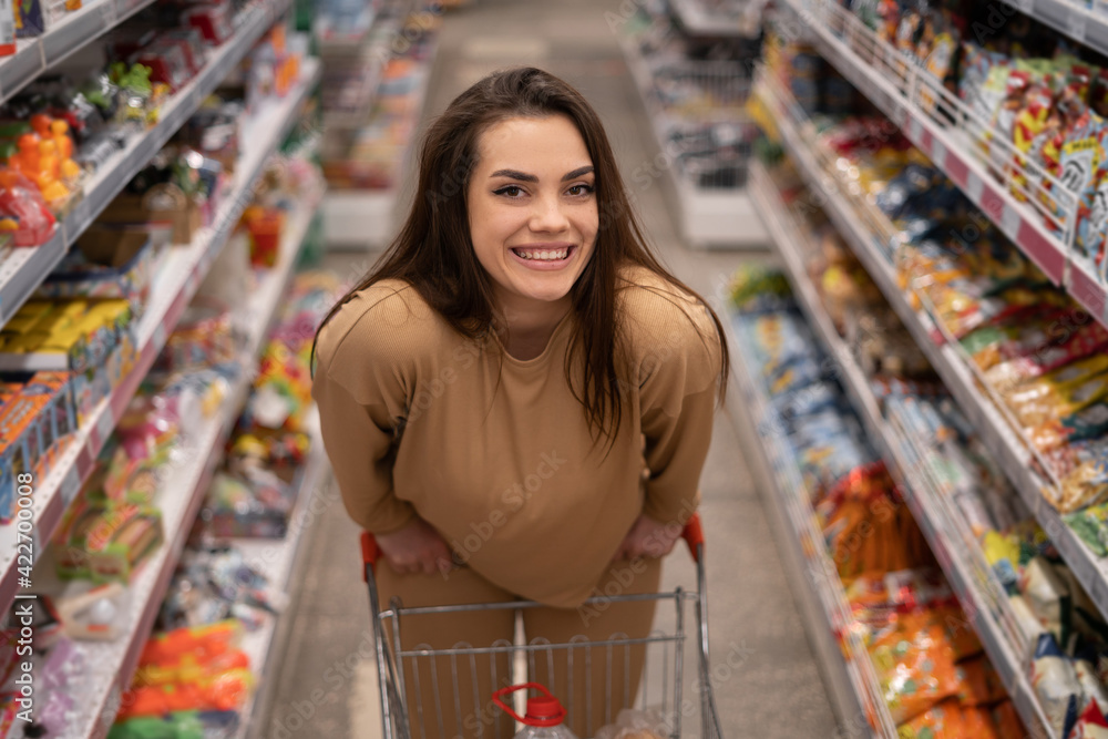 female Caucasian customer posing with shopping cart buying food in supermarket looking at camera. brunette choosing products in the store.