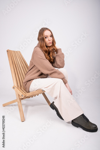 Beautiful woman with curly red hair in beige pants and a taupe hoodie sitting in wooden armchair on studio background.