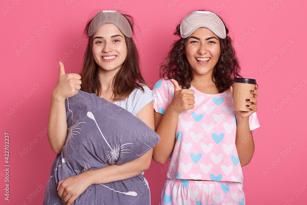 happy european women standing with satisfied facial expressions, having fun together, posing in sleeping mask and pajamas, holding coffee in disposable cup, show thumb up, wake up with good mood. happy european women standing with satisfied facial expressions, having fun together, posing in sleeping mask and pajamas, holding coffee in disposable cup, show thumb up, wake up with good mood.