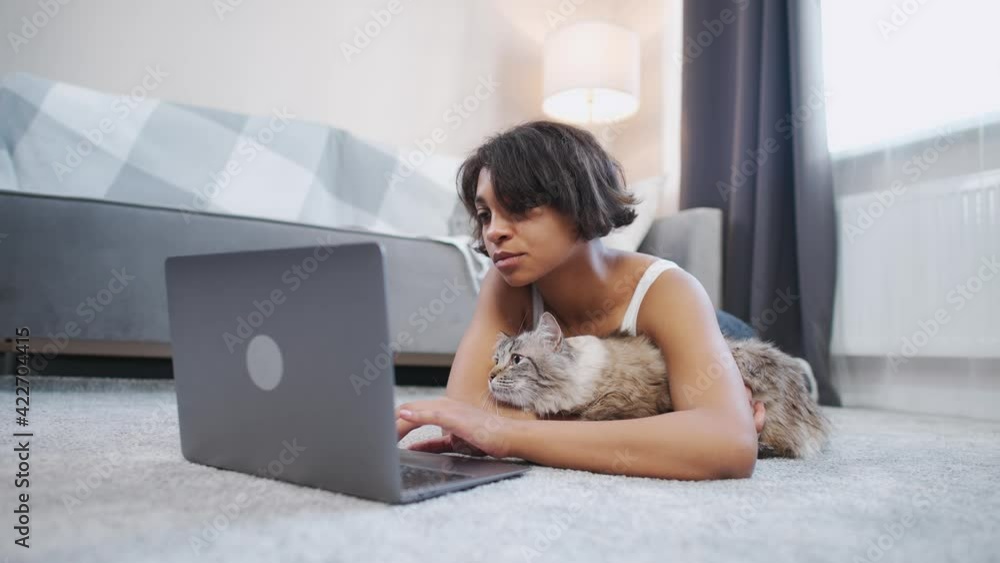 Young female with a cat lies on the floor in the living room and works at a laptop, self-isolation at home during quarantine, remote work. Young female with a cat lies on the floor in the living room and works at a laptop, self-isolation at home during quarantine, remote work.
