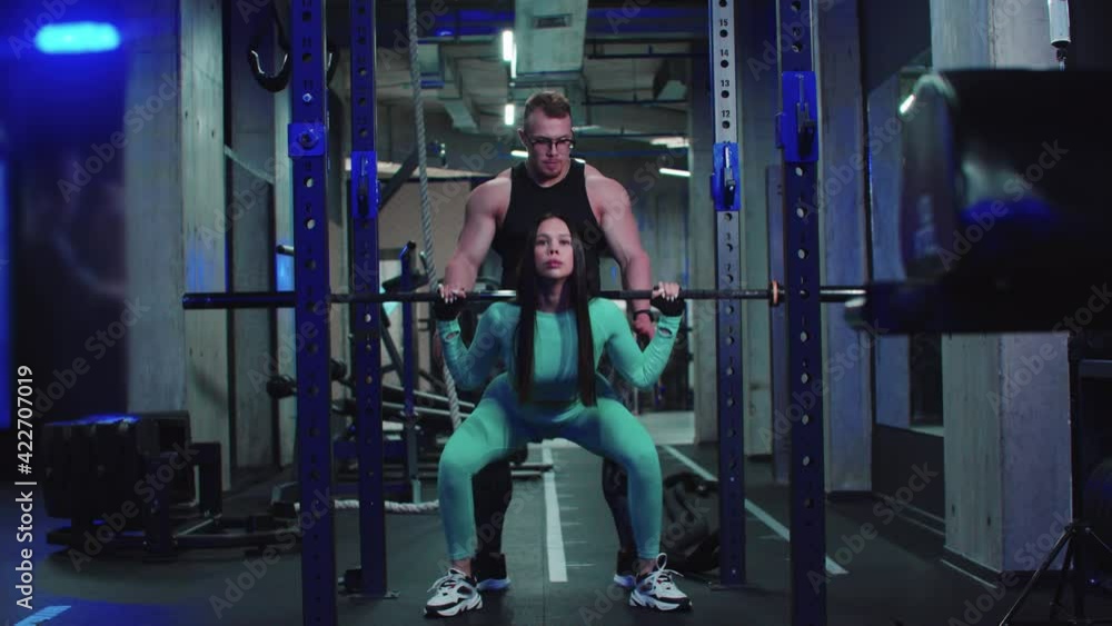Woman in sports costume trains her butt with dumbbell on her shoulders under the supervision of a trainer Woman in sports costume trains her butt with dumbbell on her shoulders under the supervision of a trainer