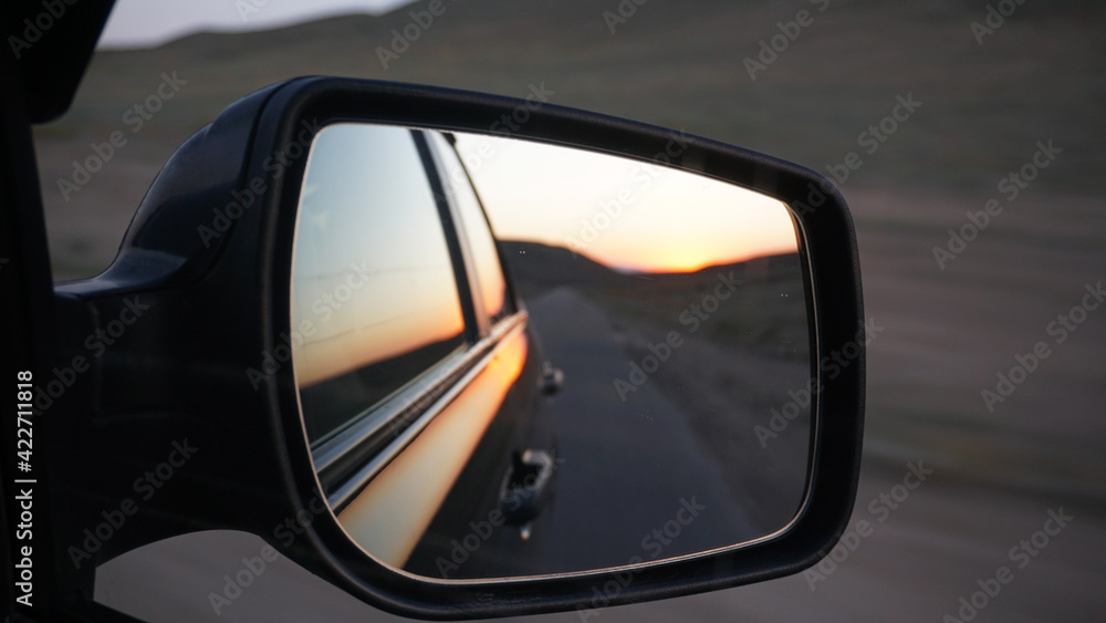 View in the side mirror of the car. Orange dawn over the hills. The car is going at high speed. Green fields, grass, and meadows are visible. Black color of the car.