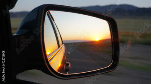 View in the side mirror of the car. Orange dawn over the hills. The car is going at high speed. Green fields, grass, and meadows are visible. Black color of the car. © SergeyPanikhin