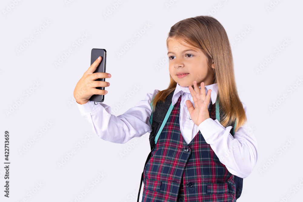 Distance learning during covid 19. A baby girl in school uniform waves her hand in front of a smartphone camera as a sign of greeting. White background. Distance learning during covid 19. A baby girl in school uniform waves her hand in front of a smartphone camera as a sign of greeting. White background.