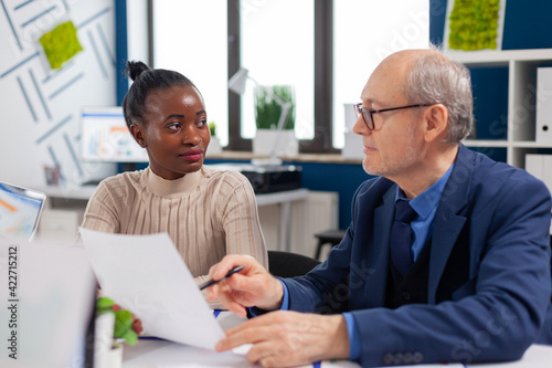 African employee discussing with senior executive looking financial charts in startup company conference board room. Multiethnic businesswoman and mature entrepreneur brainstorming.