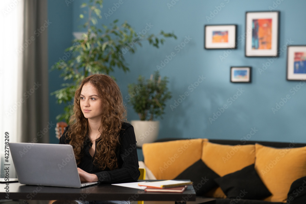 Fototapeta premium A young college girl is wondering what to write back to an email she received. The girl is sitting at a desk in a cozy living room using her laptop. The smiling teenage girl is studying remotely.