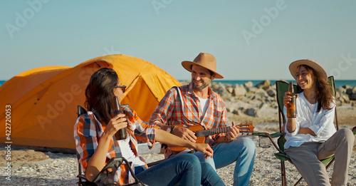 friends relaxing on beach. handsome man in hat playing ukulele and two beautiful girlfriends getting fun at seaside