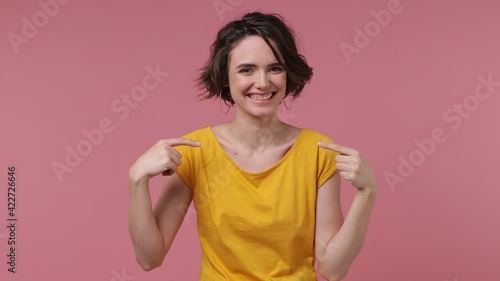 Confident brunette short hairdo young woman 20s wears basic casual yellow t-shirt looking camera charming smile pointing fingers on herself blinking isolated on pastel pink background studio portrait