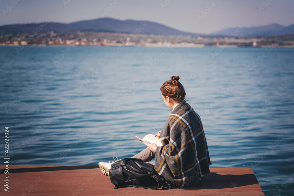 Fototapeta premium A young woman artist sitting on a pier on a sunny day and draws a landscape in a sketchbook