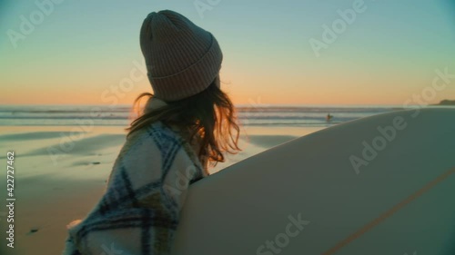 Handheld cinematic shot of urban millennial young woman walk with surfboard under arm on epic beautiful beach at sunset, after evening session of surfing. California vibes lifestyle photo