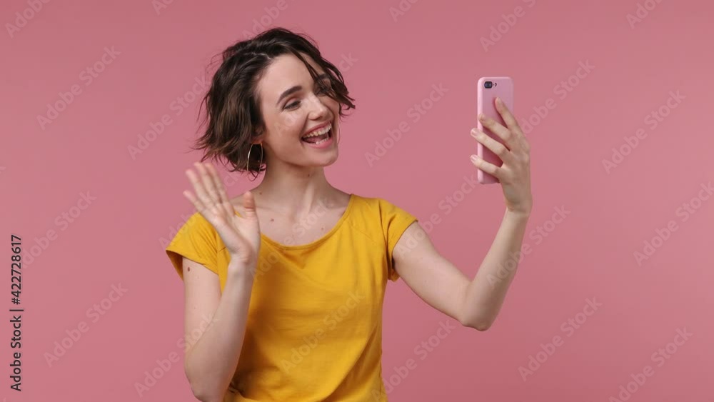 Young woman 20s wears yellow t-shirt get video call using mobile cell phone doing selfie videoconference talk conducting pleasant conversation greet with hand isolated on pastel pink background studio Young woman 20s wears yellow t-shirt get video call using mobile cell phone doing selfie videoconference talk conducting pleasant conversation greet with hand isolated on pastel pink background studio