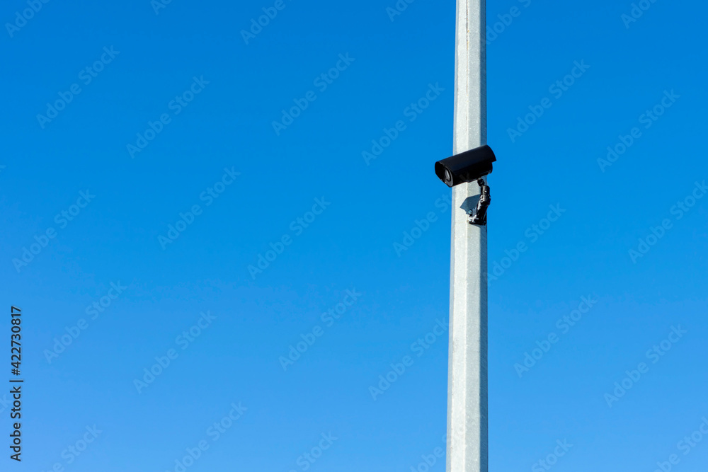 Black CCTV camera on gray pole against blue sky. Sunny winter day. Side view. Black CCTV camera on gray pole against blue sky. Sunny winter day. Side view.
