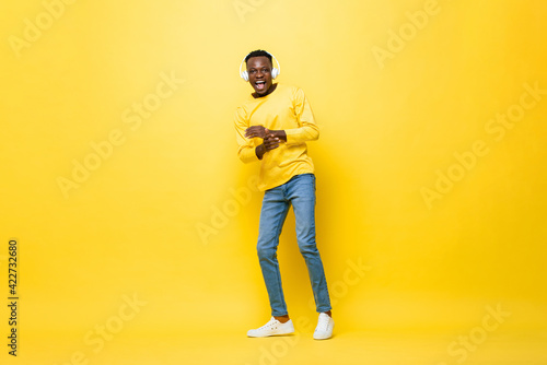 Fototapeta Naklejka Na Ścianę i Meble -  Young happy African man wearing headphones listening to music and dancing in isolated yellow studio background