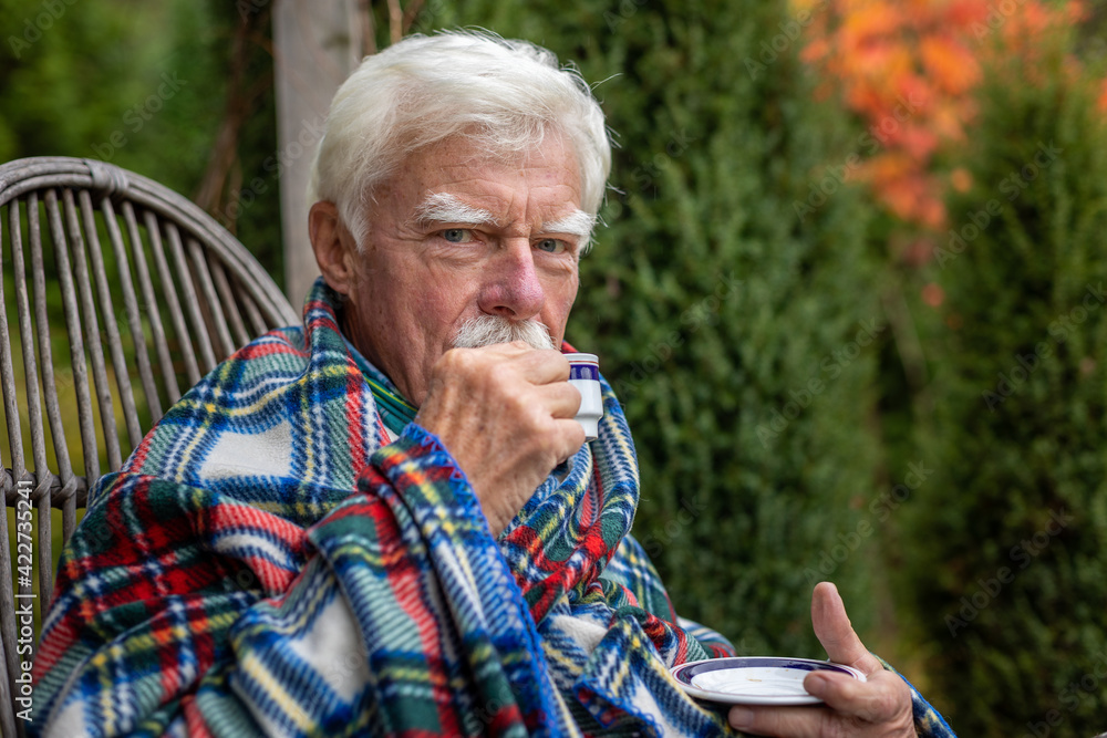 An older man wrapped in a blanket relaxes on a porch in the garden, drinking coffee. An older man wrapped in a blanket relaxes on a porch in the garden, drinking coffee.
