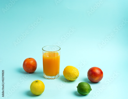 A glass of orange juice surrounded by limes, lemons and oranges on a blue background.
