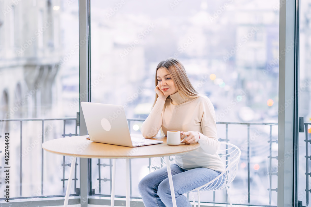 Concentrated young business woman or student looking at laptop holding coffee cup studying, serious woman working or studying with computer doing research or preparing for exam online Concentrated young business woman or student looking at laptop holding coffee cup studying, serious woman working or studying with computer doing research or preparing for exam online