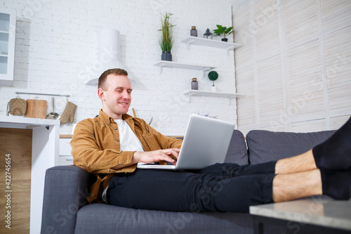 A young successful man is sitting at home on the couch with a laptop and working. Remote work during quarantine. © Дмитрий Ткачук