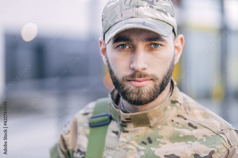a serious young soldier standing in the street, wearing a cap and special clothing in green color.