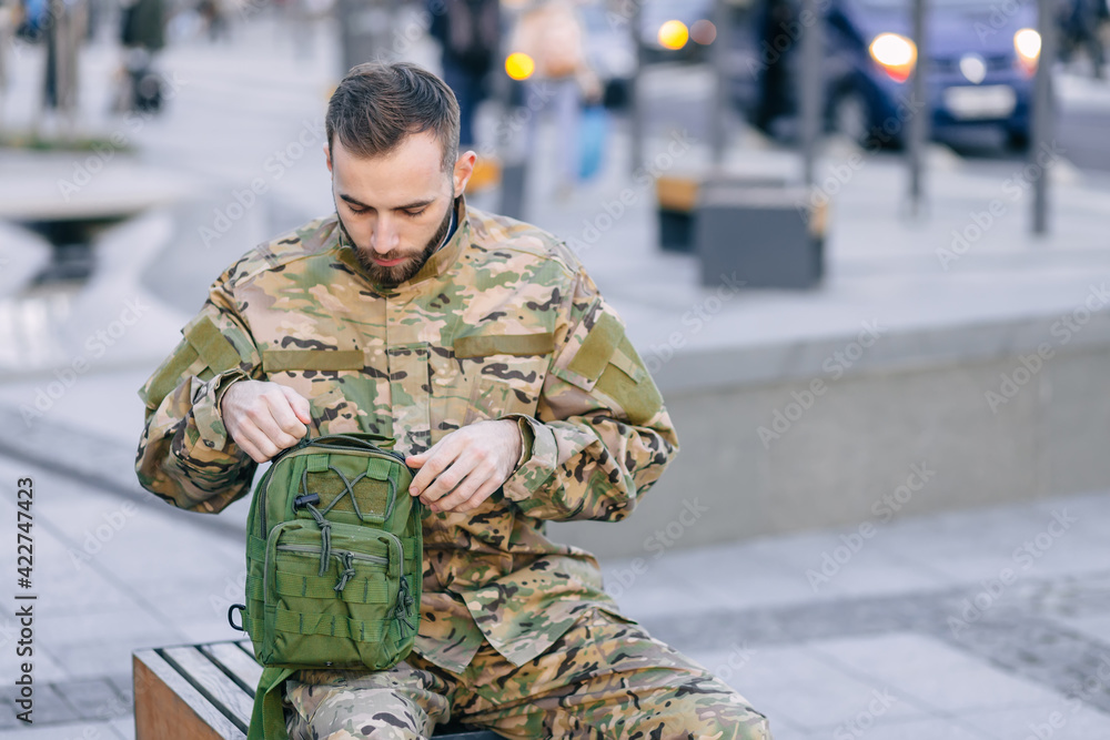 a young soldier in camouflage uniform sits on a bench, checks his bag, heading home after a long service a young soldier in camouflage uniform sits on a bench, checks his bag, heading home after a long service