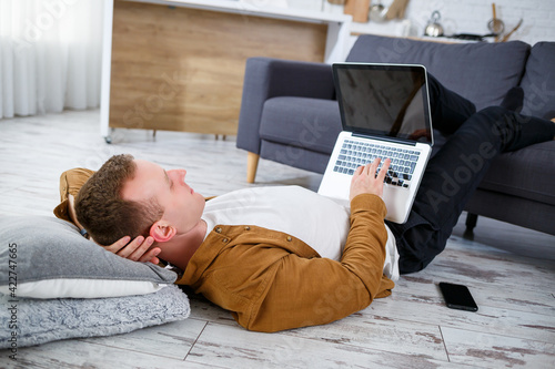Attractive smiling young man in casual wear sitting on floor on sofa in living room using laptop computer © Дмитрий Ткачук