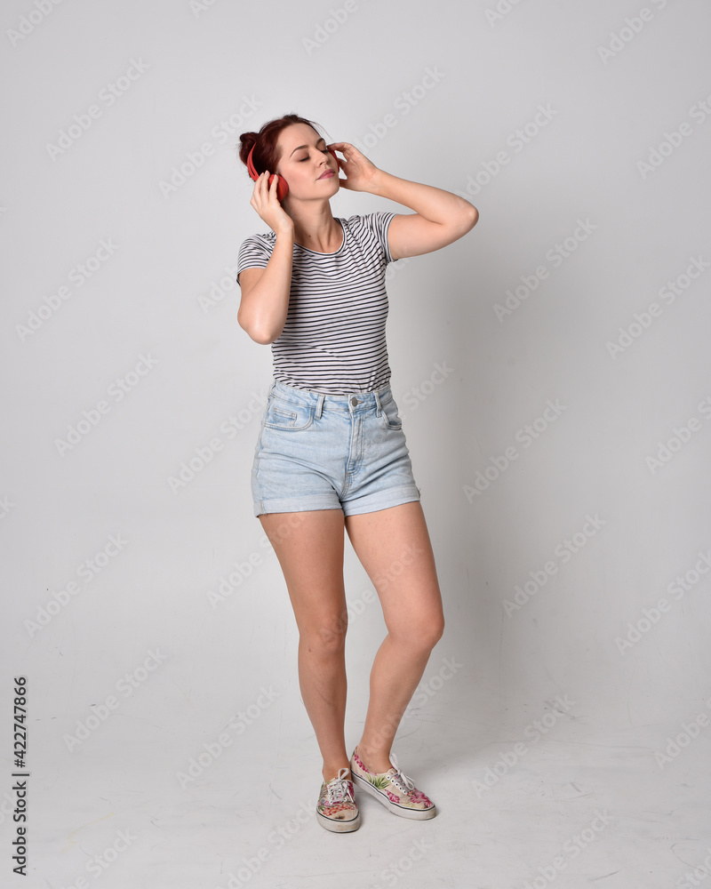 full length portrait of girl wearing shirt and denim shorts and headphones. isolated on grey studio background. full length portrait of girl wearing shirt and denim shorts and headphones. isolated on grey studio background.