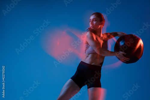 Caucasian professional female athlete training on blue studio background in neon, mixed light. Muscular, sportive woman.