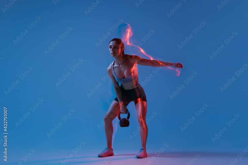 Caucasian professional female athlete training on blue studio background in neon, mixed light. Muscular, sportive woman.