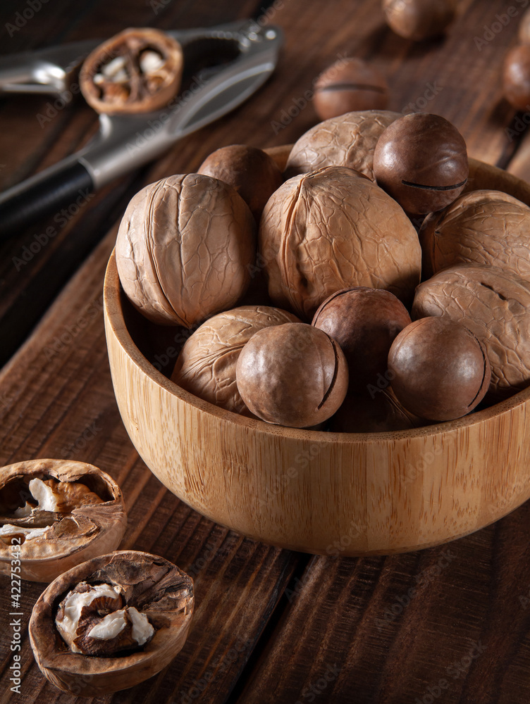 Obraz premium Walnuts and macadamia nuts in a bamboo bowl on wooden table with nutcracker in the background, vertical, close-up
