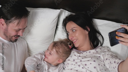 Happy young family with a baby girl, are photographed on a smartphone in the bed. Mother, father and daughter-bloggers in the room communicate using a modern gadget online. Modern young family