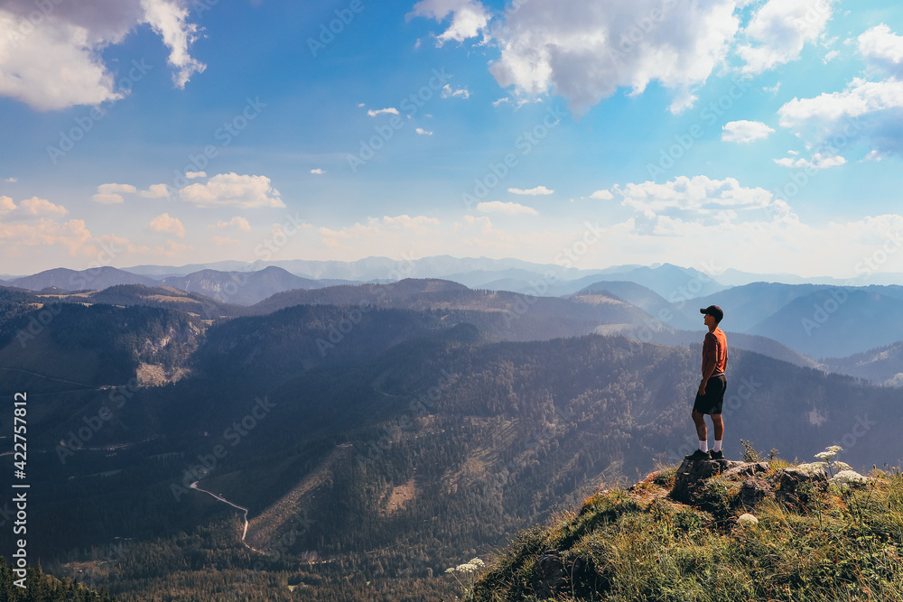 Charging energy in wild Austrian nature. Athlete of lighter figure with cap and red elastic trick stands on the edge of rock and enjoys feeling of relaxation and freedom. Conquering Mount Otscher