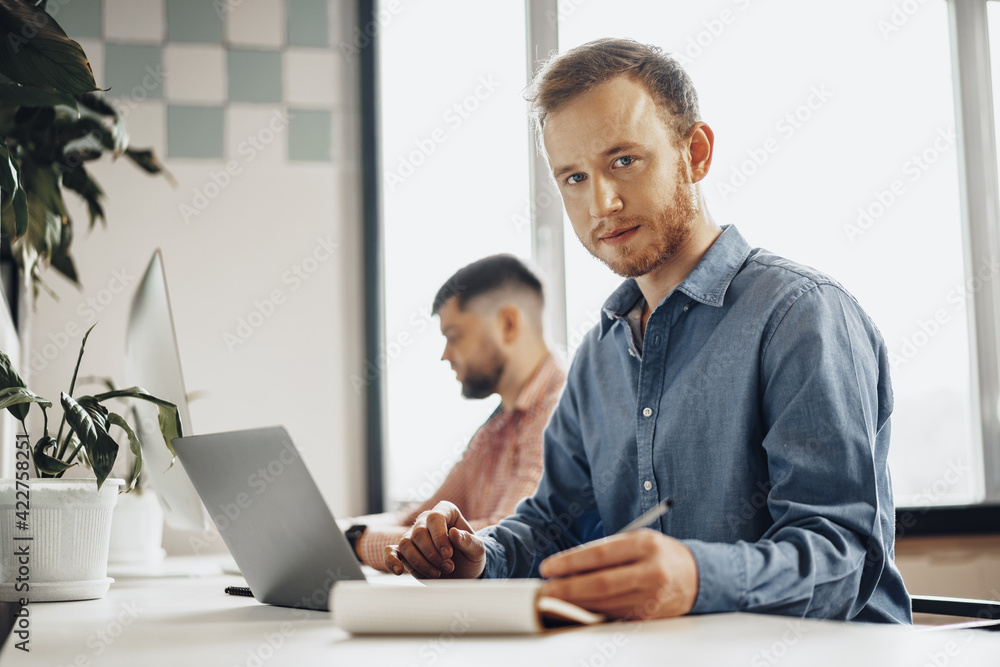 Young businessman making notes in notepad while sitting at the working table in modern office Young businessman making notes in notepad while sitting at the working table in modern office