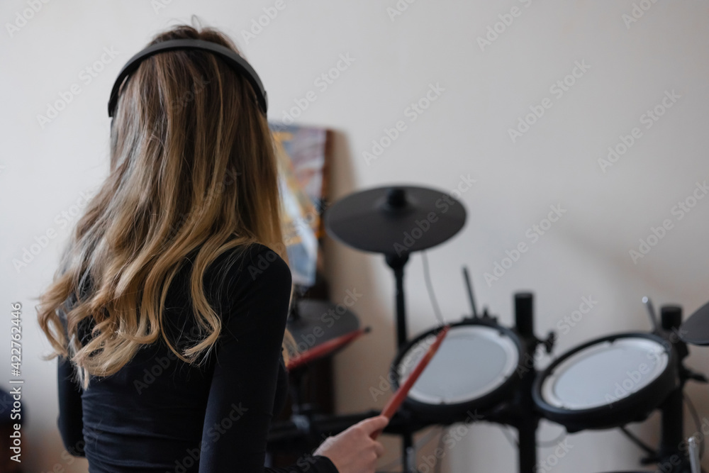 A beautiful girl in headphones sits at an electronic drum kit in the studio. Plays with drumsticks. View from the back. Blonde curly hair
