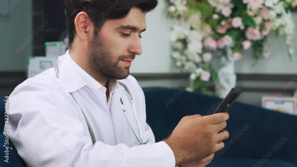 Portrait of male doctor in the lobby at modern clinic. Looking at smartphone. Wearing lab coat and stethoscope sitting on sofa. Relaxation after work. Healthcare app on cell technology Portrait of male doctor in the lobby at modern clinic. Looking at smartphone. Wearing lab coat and stethoscope sitting on sofa. Relaxation after work. Healthcare app on cell technology
