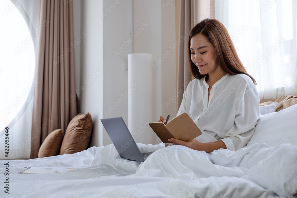 A beautiful young asian woman using laptop computer and working on paperwork while sitting on bed at home A beautiful young asian woman using laptop computer and working on paperwork while sitting on bed at home