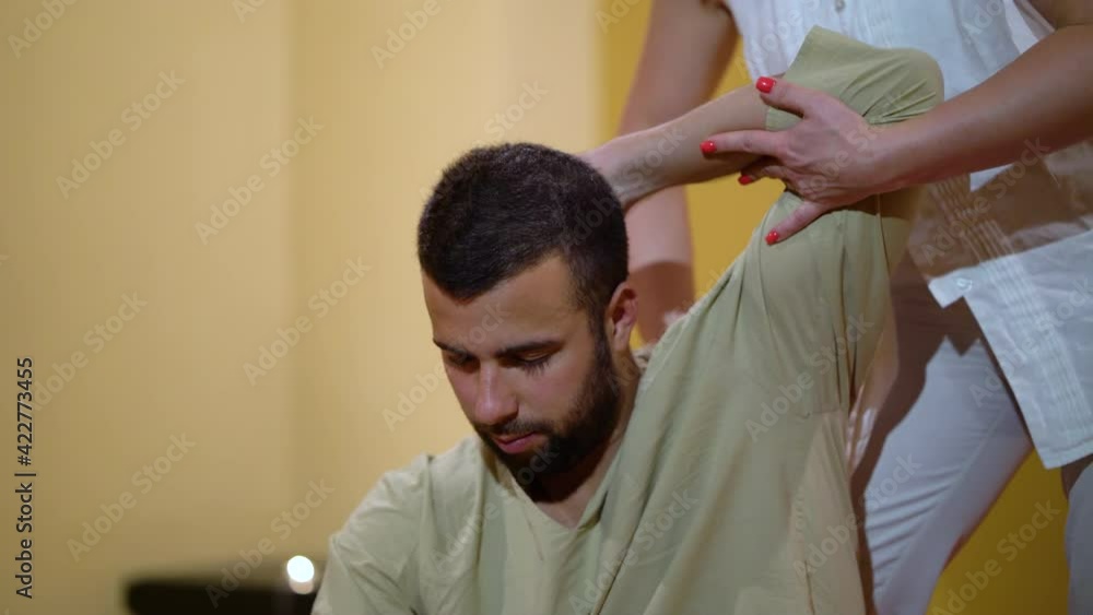 Bearded man sitting with closed eyes and getting traditional Thai massage. Handheld female therapist in white uniform standing behind and pulling his arm and shoulder. Concept of body care Bearded man sitting with closed eyes and getting traditional Thai massage. Handheld female therapist in white uniform standing behind and pulling his arm and shoulder. Concept of body care