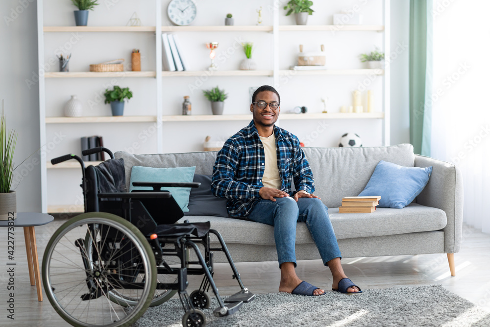 Happy disabled black guy sitting on sofa with books, empty wheelchair standing nearby, full length portrait Happy disabled black guy sitting on sofa with books, empty wheelchair standing nearby, full length portrait