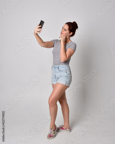 full length portrait of girl wearing shirt and denim shorts. Standing pose holding a phone isolated on grey studio background.