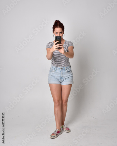 full length portrait of girl wearing shirt and denim shorts. Standing pose holding a phone isolated on grey studio background.