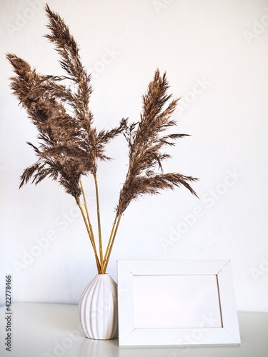 White frame mock up and dried natural pampas grass in white vase on bookshelf or table. Interior decoration element. Background boho. Minimalism design. Place for text. Selective focuson photo