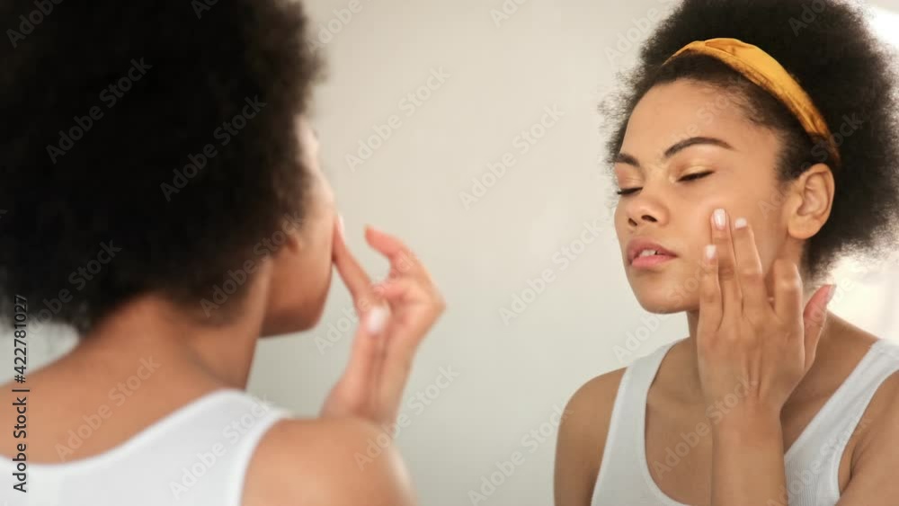 Black african american woman applying moisturizing cream on her face daily beauty skin care cosmetic routine in front a mirror at home. Black african american woman applying moisturizing cream on her face daily beauty skin care cosmetic routine in front a mirror at home.