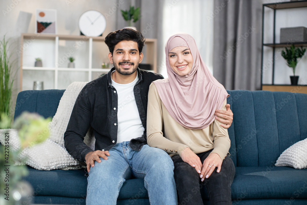 Portrait of happy young muslim couple sitting together at home on blue soft couch at cozy living room, smiling to camera. Portrait of happy young muslim couple sitting together at home on blue soft couch at cozy living room, smiling to camera.