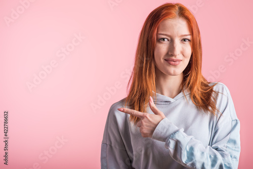 Redhead girl showing index finger to the side for copyspace in studio on pink background