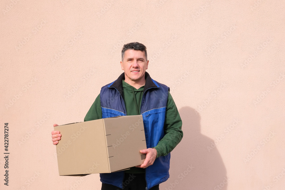 Middle-aged delivery man in uniform holding and carrying a cardbox on pink wall background. Volunteer delivery of food or medicine during the coronavirus pandemic Middle-aged delivery man in uniform holding and carrying a cardbox on pink wall background. Volunteer delivery of food or medicine during the coronavirus pandemic